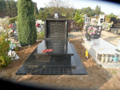 Czarny, polerowany nagrobek z granitu na cmentarzu, otoczony piaskiem. Widoczne inskrypcje i dekoracje, w tle inne groby i drzewa. Kadr z bliska, oświetlenie dzienne.