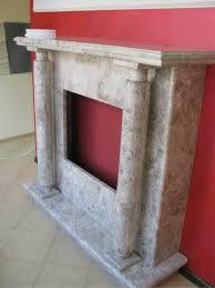 Marmurowy kominek z kolumnami na jasnej podłodze, na tle czerwonej ściany. Widoczne detale wykończenia i struktura kamienia. Dekoracyjny element wnętrza.
