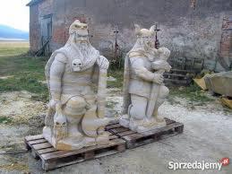 Dwie rzeźby ogrodowe przedstawiające brodatych wojowników w hełmach, ustawione na drewnianych paletach. Tło stanowi ceglany budynek i zielone pole.