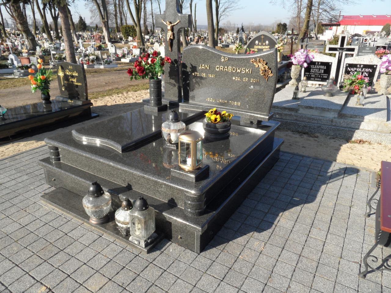 Czarny, granitowy nagrobek z inskrypcją 'Jan Grabowski' na cmentarzu, otoczony lampionami i kwiatami, w tle inne groby i drzewa. Ujęcie w słoneczny dzień.