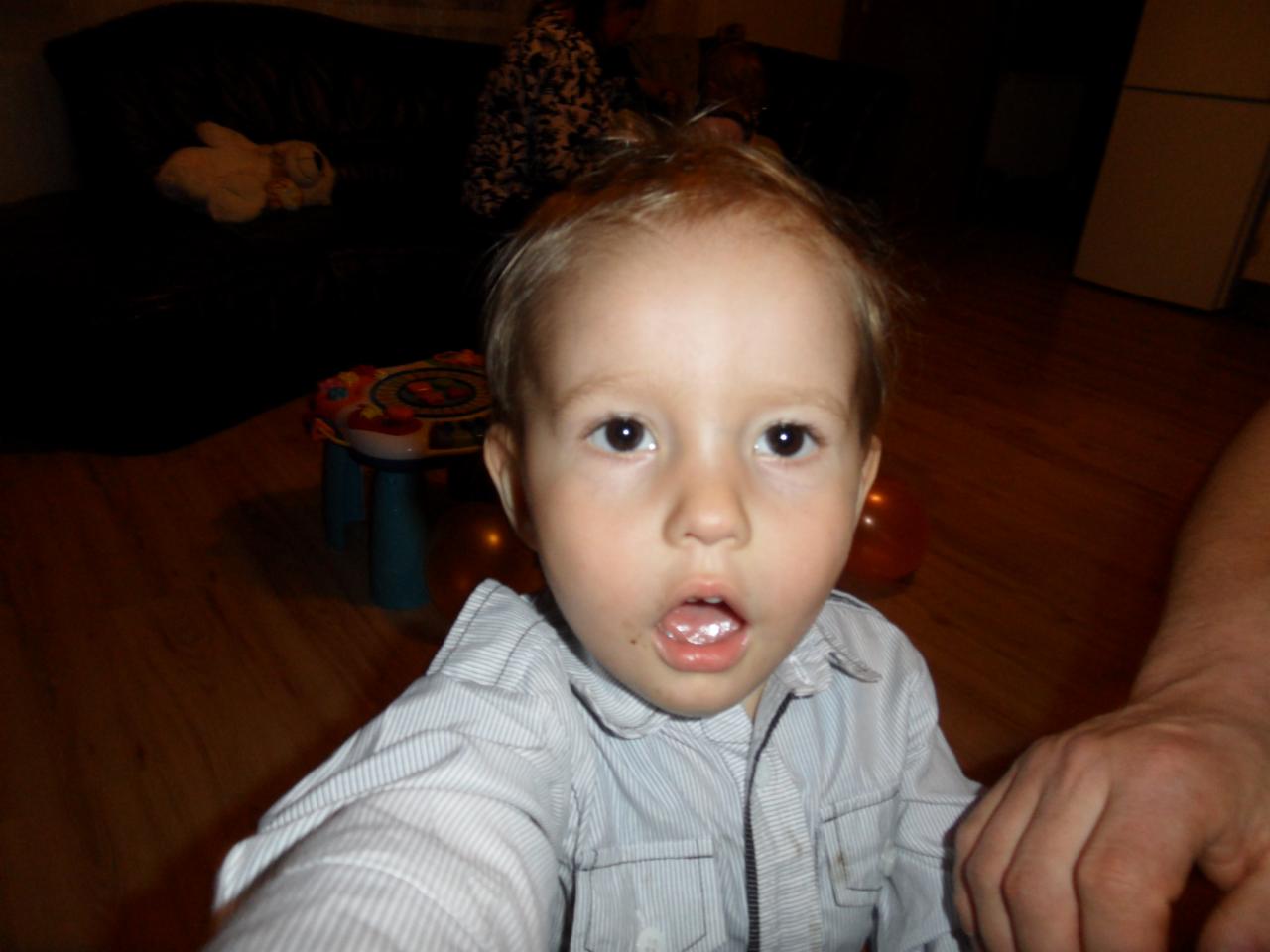 Zbliżenie na twarz otwartego na świat, zaskoczonego chłopca w szarej koszuli na tle pokoju z drewnianą podłogą i zabawkami, z widoczną dłonią dorosłego.