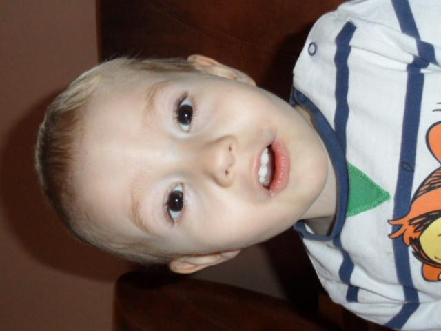 Portret uśmiechniętego blond chłopca w koszulce w paski z postacią z kreskówki, skierowany do góry nogami, z widocznymi pierwszymi zębami.