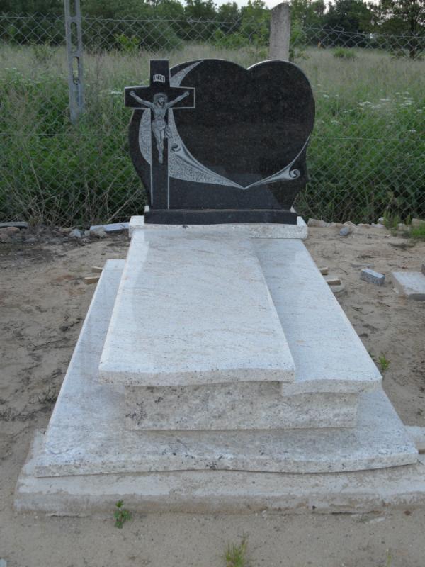 Czarny nagrobek z krzyżem i sercem na jasnym, granitowym cokole. Pomnik stoi na piasku, w tle widoczna zieleń i ogrodzenie z siatki.