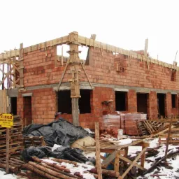 Wznoszenie dwukondygnacyjnego budynku z cegły klinkierowej. Widoczne drewniane szalunki i rusztowania. Na pierwszym planie palety i materiały budowlane.