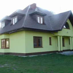 Żółty dom jednorodzinny z brązowym dachem i oknami, widok z zewnątrz. Dachówka ceramiczna, widoczne rynny i podbitka. Elewacja w odcieniach żółci z brązowym cokołem.