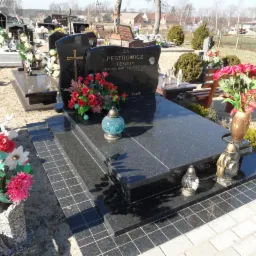 Czarny, polerowany nagrobek z inskrypcją i kwiatami, otoczony innymi nagrobkami na cmentarzu w słoneczny dzień. Widoczne schodki z płytek prowadzące do pomnika.