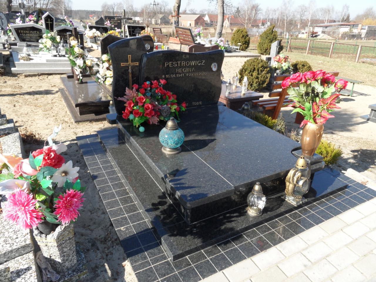 Czarny, polerowany nagrobek z inskrypcją i kwiatami, otoczony innymi nagrobkami na cmentarzu w słoneczny dzień. Widoczne schodki z płytek prowadzące do pomnika.