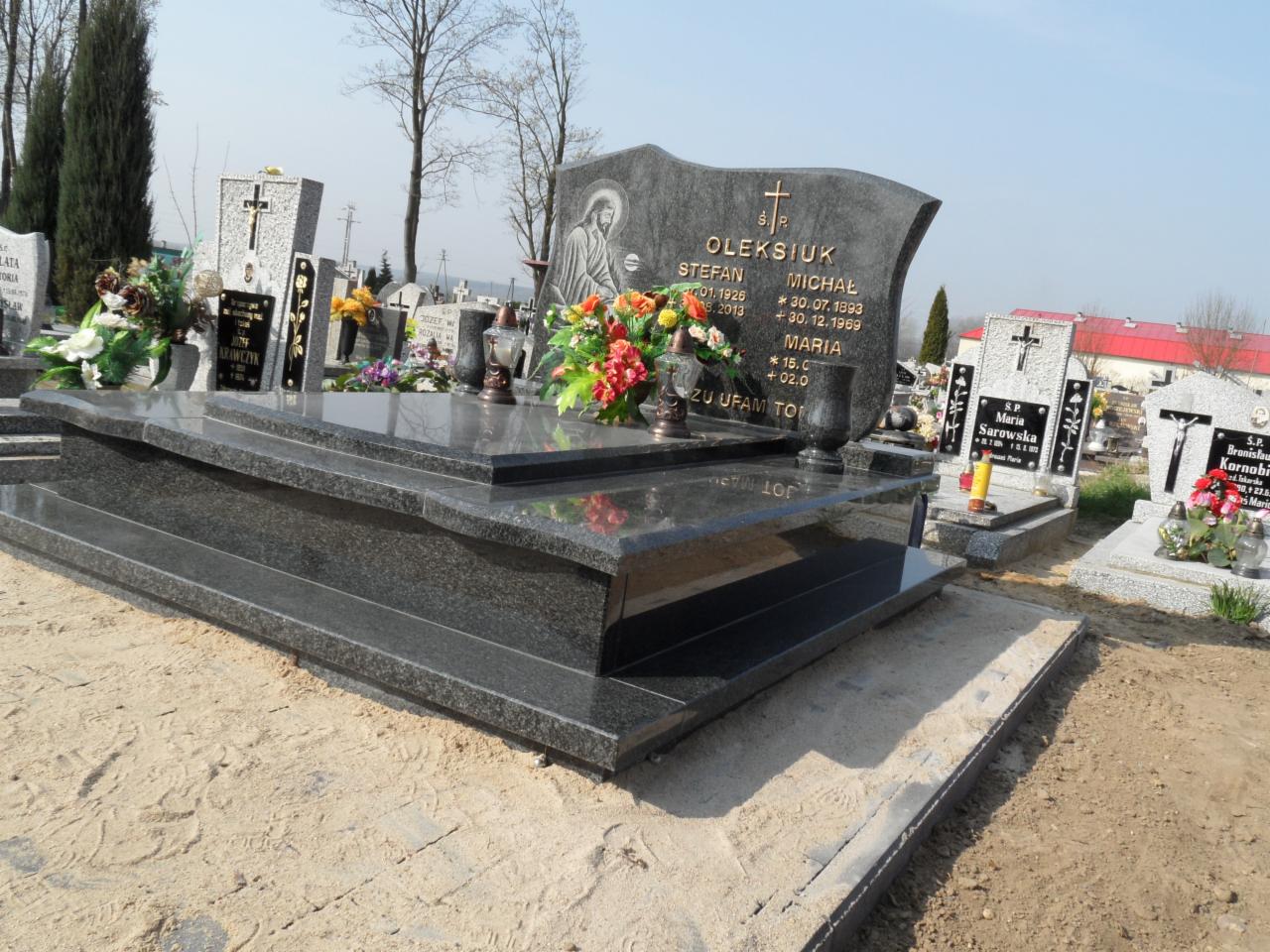 Granitowy nagrobek z inskrypcjami i kwiatami na cmentarzu, w tle inne nagrobki i drzewa. Jasne, dzienne światło.