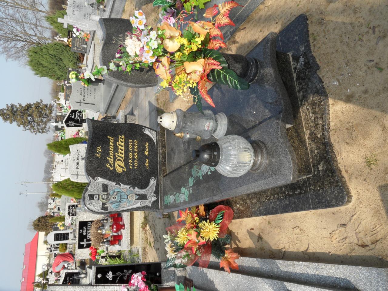 Czarny nagrobek z inskrypcjami i zdobieniami, ozdobiony kwiatami i zniczami, na tle cmentarza z innymi nagrobkami i zielenią.