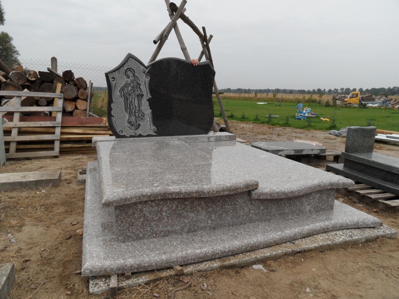 Nowy, jasnoszary nagrobek z ciemnym, grawerowanym elementem, ustawiony na placu kamieniarskim. W tle widoczne drzewa i pola uprawne pod pochmurnym niebem.