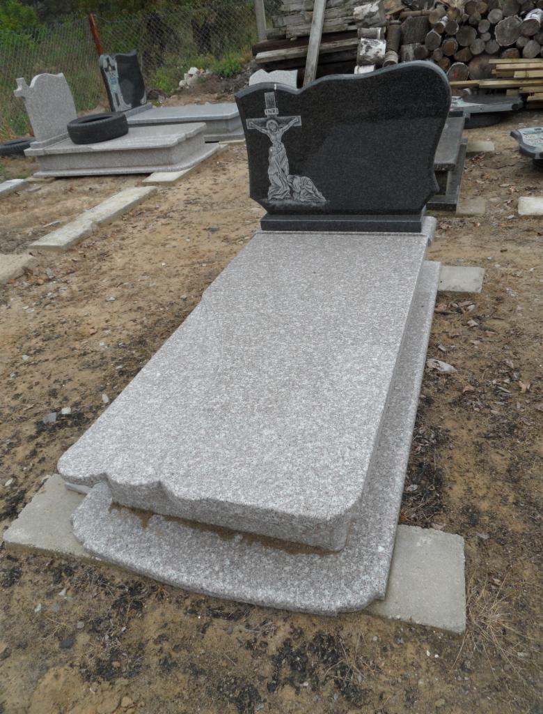 Szary, granitowy nagrobek z czarną płytą z wizerunkiem ukrzyżowanego Jezusa, ustawiony na ziemi. W tle inne nagrobki i stos drewna.