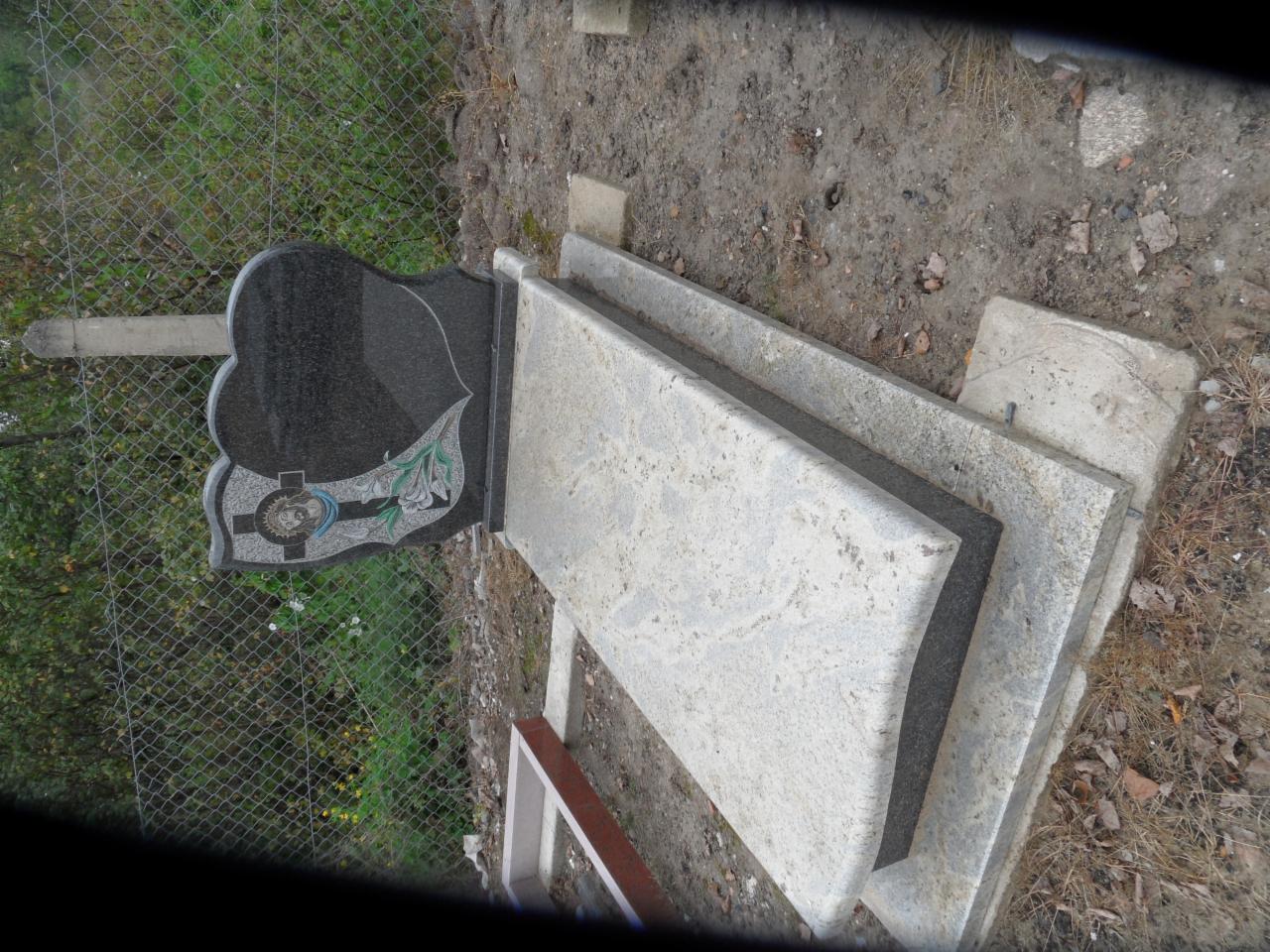 Czarny nagrobek z motywem kwiatowym i wizerunkiem religijnym, oparty o jasnoszare płyty, na zewnątrz, przy siatce ogrodzeniowej. Surowy kamień.