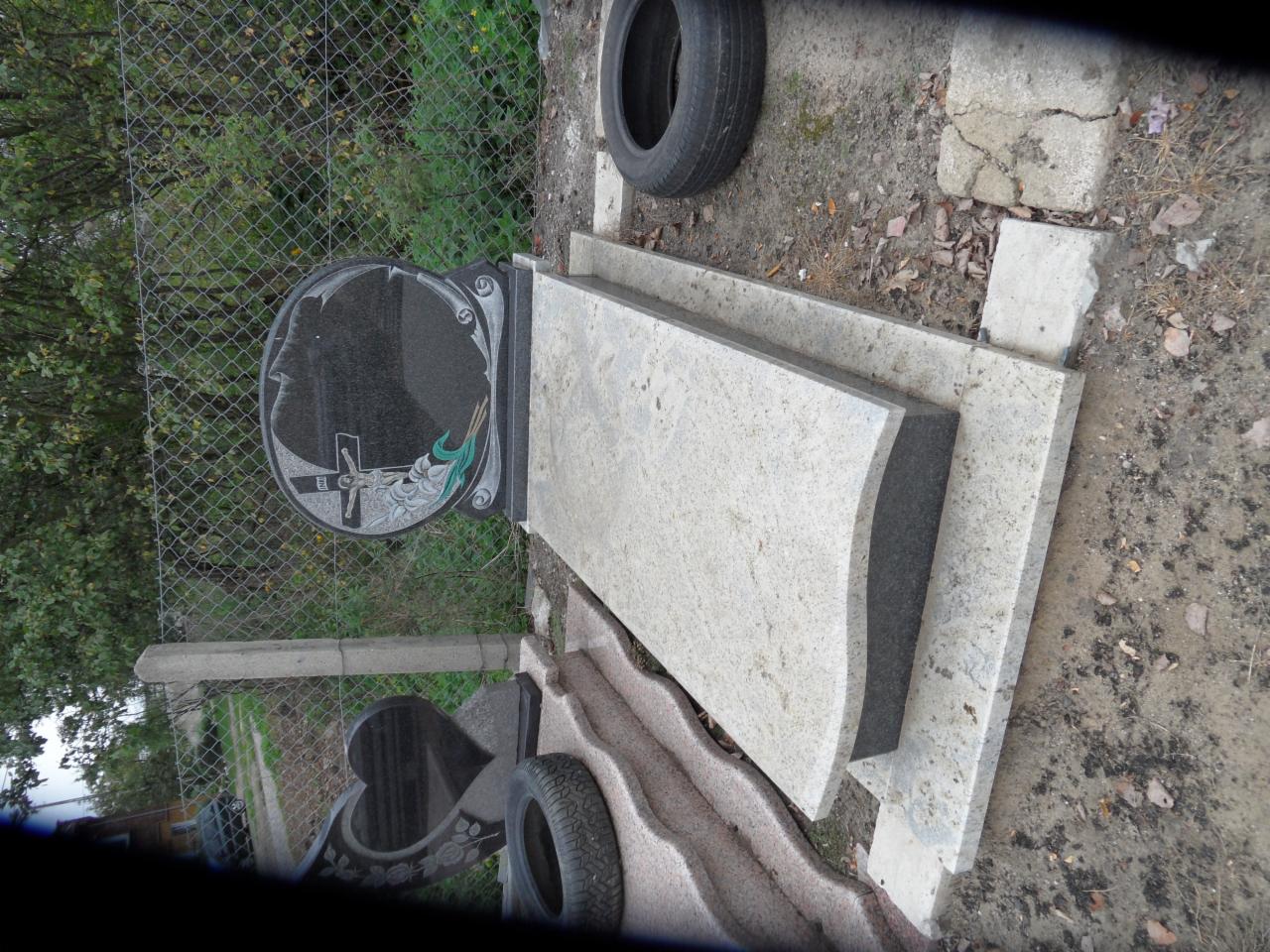 Jasny, granitowy nagrobek z czarną płytą z motywem krzyża i twarzy, ustawiony na ziemi obok opony samochodowej i ogrodzenia z siatki. Widok z boku, częściowo obrócony.