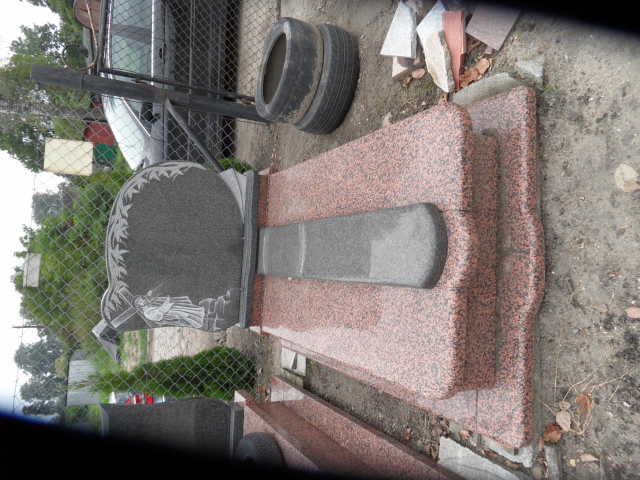 Granitowy nagrobek z rzeźbionym motywem roślinnym i postacią, ustawiony na dwupoziomowej podstawie z czerwonego granitu. W tle siatka ogrodzeniowa.