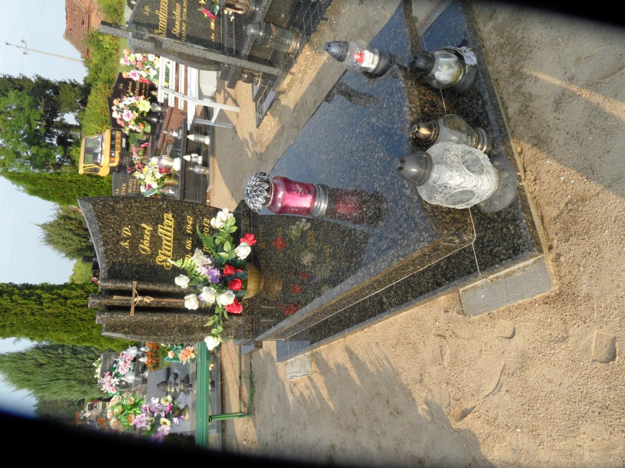 Granitowy nagrobek z krzyżem, kwiatami i lampionami na cmentarzu, widok z boku. Na płycie nagrobnej widoczne imię i nazwisko: Józef Stadler.