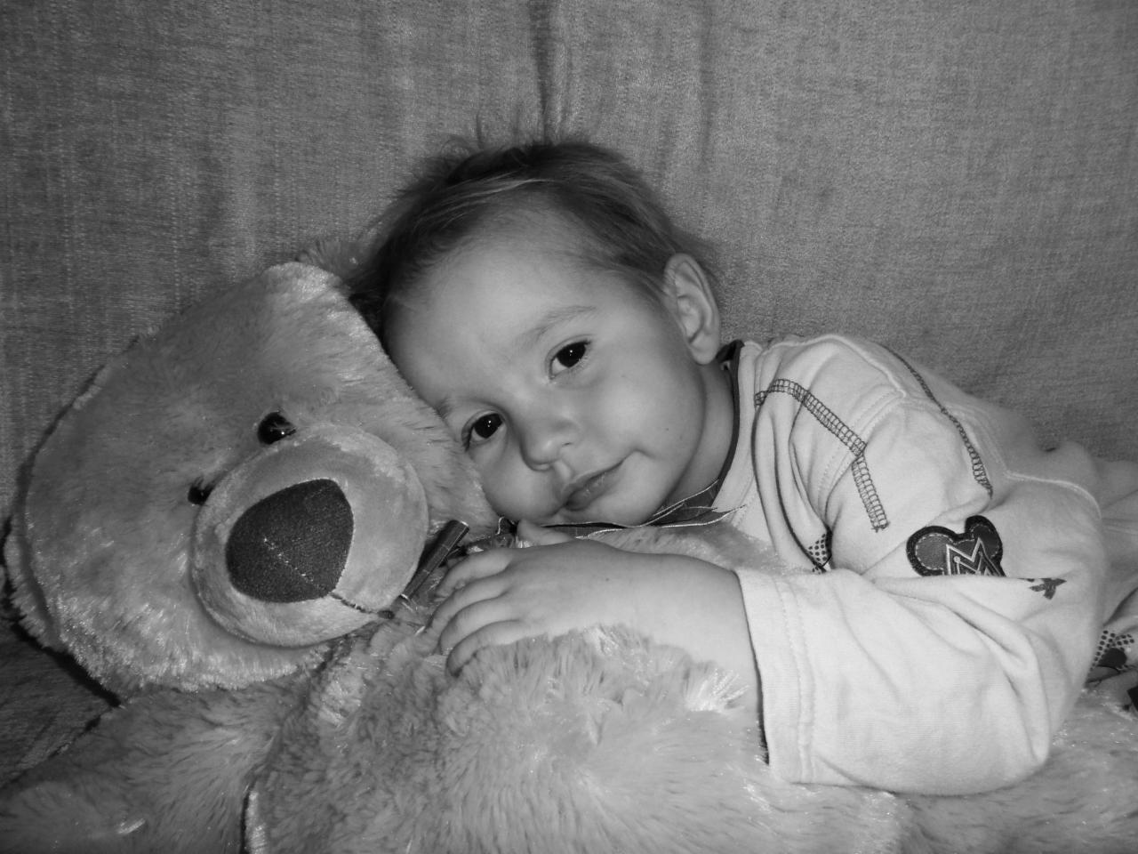 Czarno-białe zdjęcie: mały chłopiec przytula pluszowego misia, leżąc na miękkiej powierzchni, zamyślony wyraz twarzy, delikatne światło.