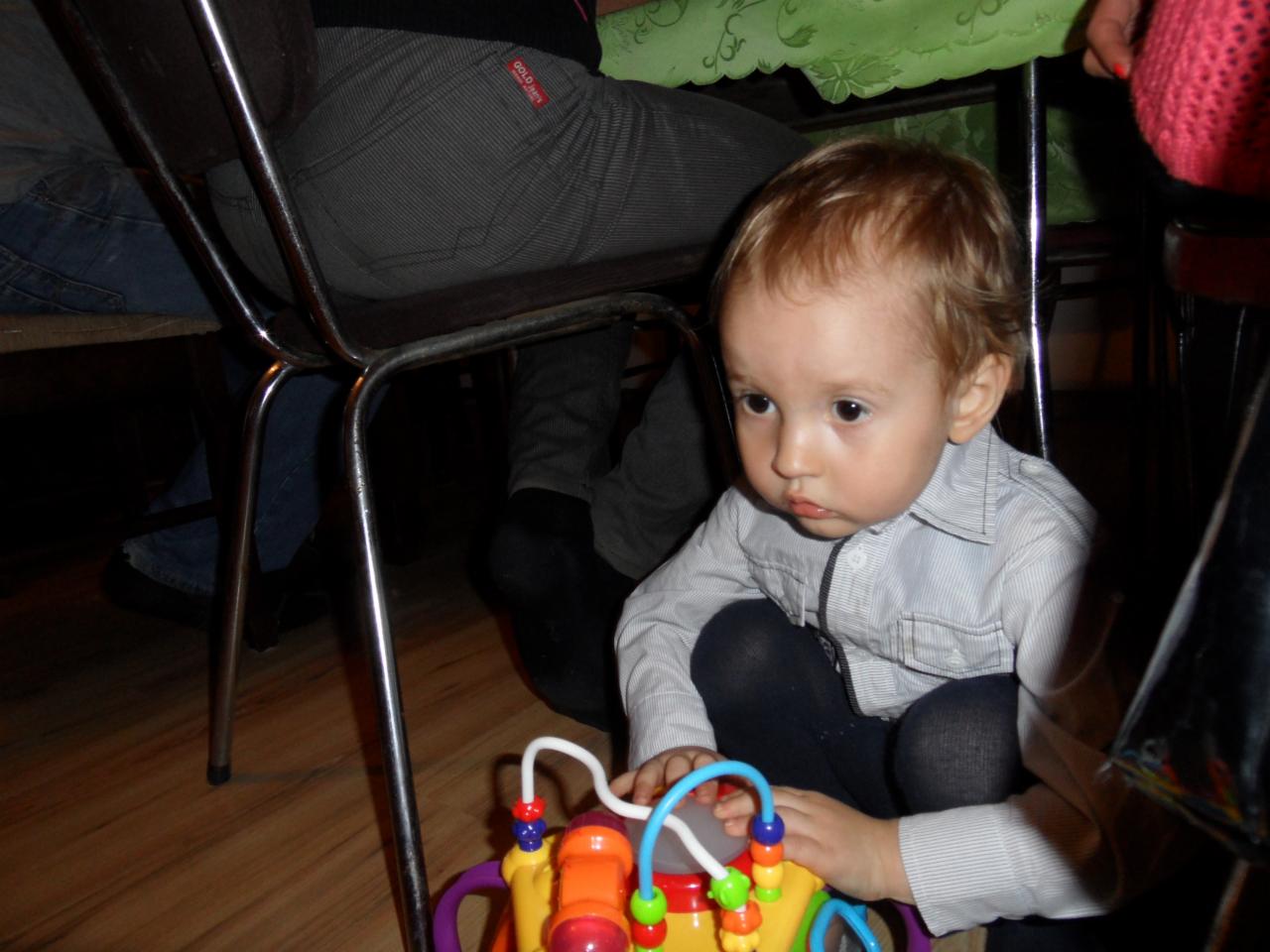 Chłopiec bawi się zabawką na drewnianej podłodze, siedząc pod stołem w otoczeniu krzeseł. Uwagę przykuwa skupiony wzrok dziecka i jego delikatna, jasna koszula.