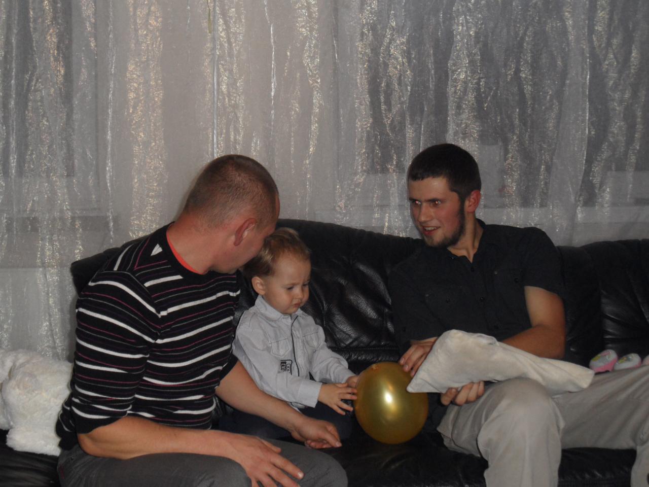 Dwóch mężczyzn i małe dziecko siedzą na czarnej skórzanej kanapie. Dziecko trzyma złoty balon. W tle firanka i okno.