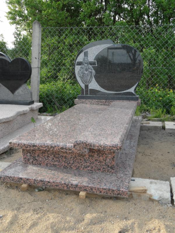 Brązowo-czarny nagrobek z rzeźbą Jezusa na tle siatki ogrodzeniowej i zieleni. Nagrobek ustawiony na drewnianych podkładkach, piaskowe podłoże.