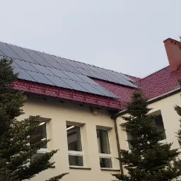 Panele słoneczne na dachu budynku z czerwoną dachówką, widoczne kominy i drzewa iglaste na pierwszym planie.