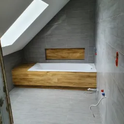 Nowoczesna łazienka na poddaszu z oknem dachowym, wykończona szarymi płytkami imitującymi kamień, z zabudowaną wanną w obudowie z drewna i widocznymi instalacjami hydraulicznymi w trakcie montażu.