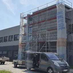 Budynek w trakcie prac elewacyjnych, rusztowanie częściowo osłonięte folią z widocznym logo producenta materiałów budowlanych, na pierwszym planie zaparkowany bus z otwartymi drzwiami i przyczepka...