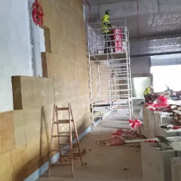 Prace ociepleniowe wewnątrz budynku: pracownik na rusztowaniu układa wełnę mineralną na ścianie z bloczków, widoczna drewniana drabina i opakowania wełny Rockwool.