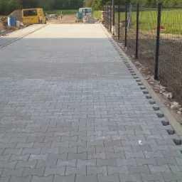 Ukończona nawierzchnia z szarej kostki brukowej typu 'I', z widocznymi obrzeżami i fragmentem ogrodzenia panelowego po prawej stronie, w tle żółty bus i przygotowany teren.