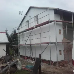 Dom w trakcie ocieplania elewacji, widoczne rusztowania, częściowo ocieplona ściana i stary mur z cegły. Na pierwszym planie bałagan i zaparkowany samochód dostawczy.