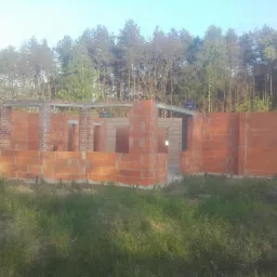 Wznoszenie ścian parterowego domu z czerwonej cegły, widoczne stalowe nadproża, w tle las