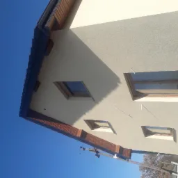 Fragment otynkowanej elewacji domu z oknami dachowymi i elementami drewnianymi pod okapem, widok pod kątem z dołu w słoneczny dzień.