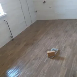 Wykończona łazienka na poddaszu z brązowymi płytkami podłogowymi imitującymi drewno i jasnymi płytkami ściennymi z dekoracyjnym pasem. Na podłodze leży gąbka i paca do fugowania.