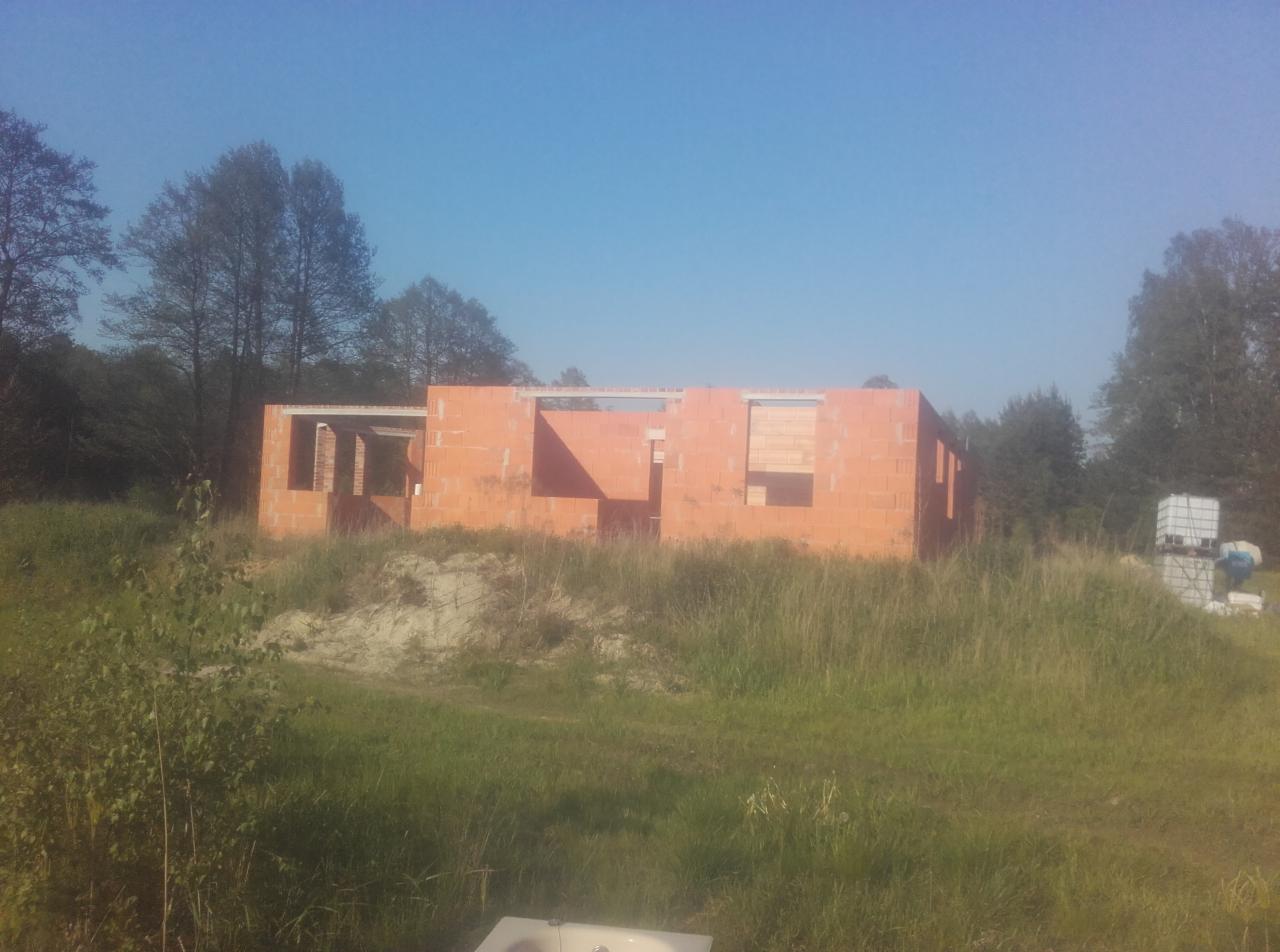 Surowa konstrukcja domu z czerwonej cegły, w otoczeniu łąki i drzew, widoczne otwory okienne i drzwiowe, z paletami materiałów w tle, w słoneczny dzień.