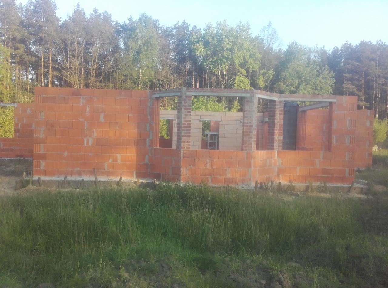 Wznoszone ściany parterowego domu z czerwonej cegły na fundamencie, widoczne otwory okienne i drzwiowe, tło stanowią drzewa.