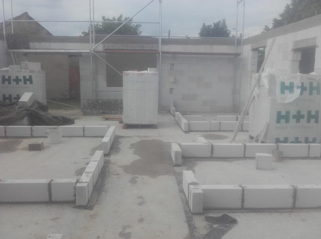 Wznoszenie ścian domu z bloczków betonowych H+H, widoczne fundamenty i początek murów, rusztowanie w tle.