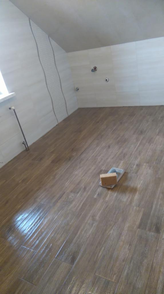 Wykończona łazienka na poddaszu z brązowymi płytkami podłogowymi imitującymi drewno i jasnymi płytkami ściennymi z dekoracyjnym pasem. Na podłodze leży gąbka i paca do fugowania.