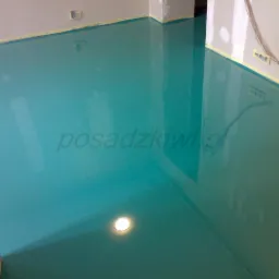 Błękitna, błyszcząca posadzka w nowym pomieszczeniu, zabezpieczona taśmą malarską przy ścianach. Widoczny punkt świetlny w podłodze.