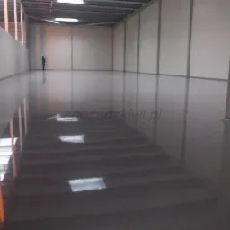 Błyszcząca, szara posadzka w dużym, jasnym magazynie. W oddali stoi osoba, co daje skale przestrzeni. Widoczne odbicia światła na powierzchni.