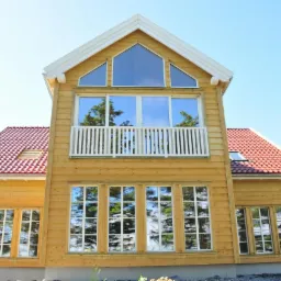 Jasny, dwukondygnacyjny dom szkieletowy z czerwonym dachem, balkonem i dużymi oknami, w których odbija się otoczenie, sfotografowany w słoneczny dzień.
