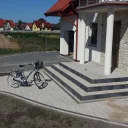 Nowoczesny podjazd z kostki brukowej prowadzący do domu z kolumnami. Na pierwszym planie rower. Widoczne domy w tle i częściowo niezagospodarowany teren.