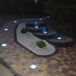 Oświetlona ścieżka z kostki brukowej i kamienny ogródek ze schodkami, nocą. Lampy LED w kostce i schodach. Dekoracyjny ogród z kruszywem i roślinami.