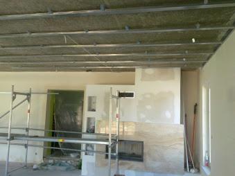 Szkielet sufitu podwieszanego z metalowych profili i izolacją, widoczne rusztowania, fragment ściany z kominkiem w trakcie prac wykończeniowych.