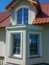 Elewacja domu z oknami w wykuszu i oknem lukarny na poddaszu. Jasna fasada, czerwona dachówka. Wykończenie zewnętrzne budynku po remoncie.