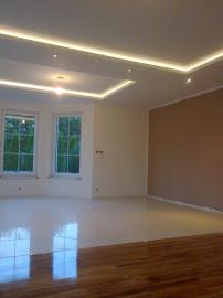 Jasne, przestronne wnętrze po remoncie: biała podłoga, drewniany parkiet, ściany w odcieniach bieli i beżu, dekoracyjne oświetlenie sufitowe LED.