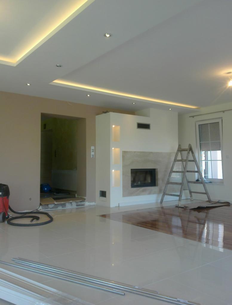 Salon po remoncie: podwieszany sufit z oświetleniem LED, kominek obłożony marmurem, błyszcząca podłoga. Widoczna drabina i odkurzacz.