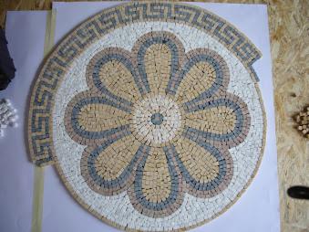 Mozaika ceramiczna w kształcie kwiatu z geometrycznym obramowaniem, ułożona na białym podkładzie. Widoczne drobne elementy mozaiki i niedokończone fragmenty.