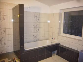Wykończona łazienka z wanną obudowaną ciemnymi płytkami, jasne płytki na ścianach z delikatnym wzorem. Widok na okno z białym obramowaniem, neutralne oświetlenie.