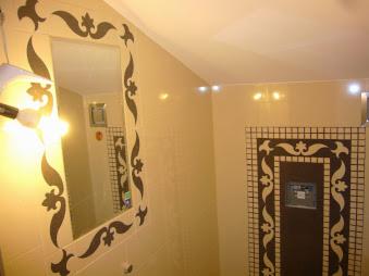 Beżowa łazienka z lustrem w dekoracyjnej ramie i mozaiką wokół drzwi rewizyjnych. Ciepłe oświetlenie podkreśla wzory na ścianach i suficie.