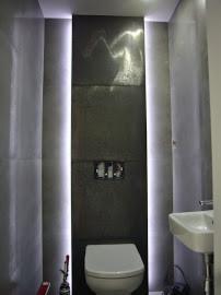 Nowoczesna toaleta: szare płytki, podświetlenie LED po bokach, minimalistyczna armatura. Widok na ścianę z zawieszonym WC i umywalką w rogu.