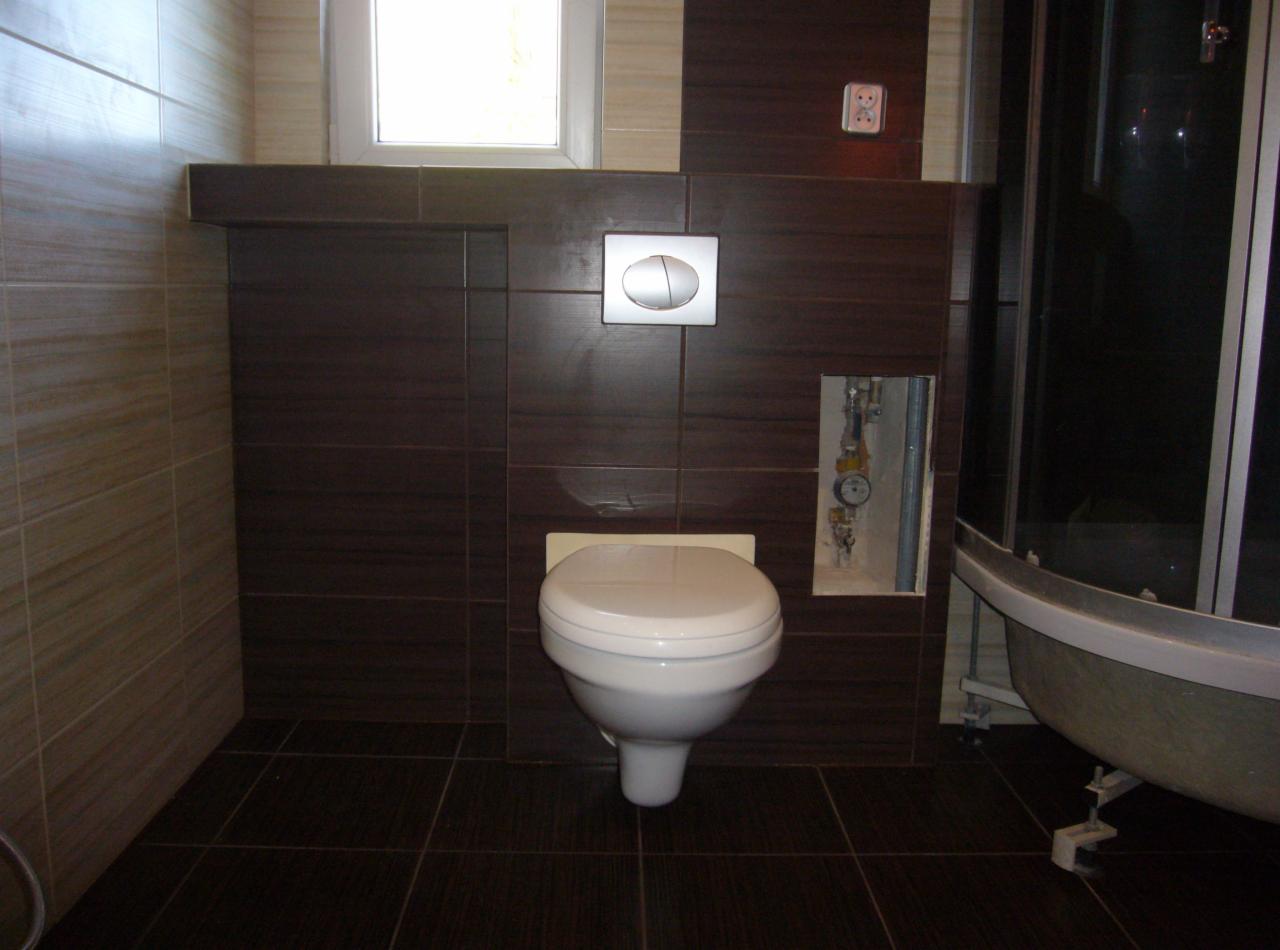 Wykończona łazienka z brązowymi płytkami, wiszącą toaletą i kabiną prysznicową. Widoczny fragment instalacji w ścianie i okno. Minimalistyczny styl.